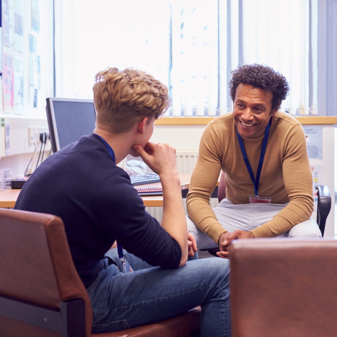 A teacher and student sit in an office, engaged in a friendly conversation. The teacher, smiling warmly, leans forward. A computer is visible in the background.
