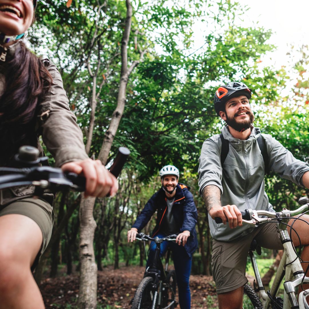 Three people riding bicycles in a lush, green forest, smiling and enjoying the outdoors. They wear casual clothing and helmets, conveying happiness.