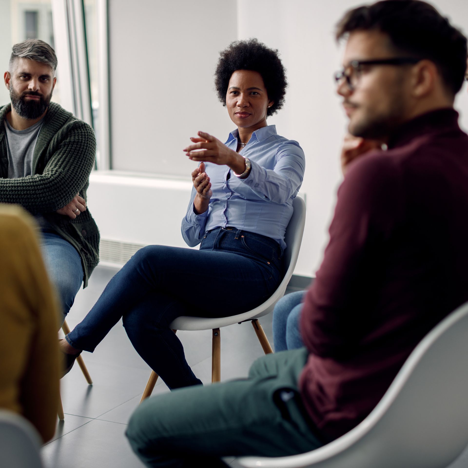 A woman in a blue shirt gestures while speaking during a group discussion in a bright room. Three other people listen intently, creating an engaged atmosphere.